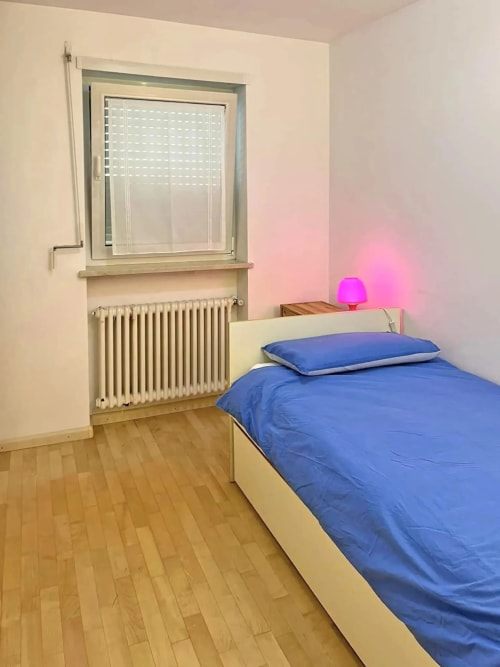 Camera luminosa nell'appartamento di Siusi con biancheria blu, pareti color crema, pavimento in legno, radiatore e lampada rosa che creano un'atmosfera accogliente