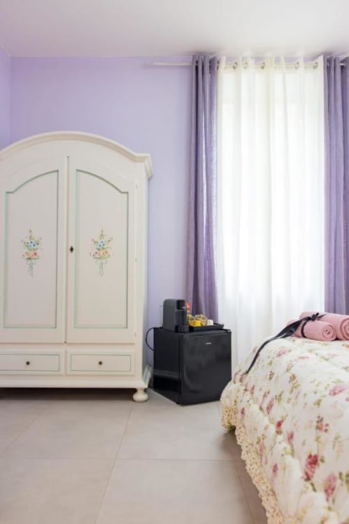Elegante camera a La Maison Du Lion con pareti lavanda, armadio color crema, biancheria floreale e tende trasparenti a La Spezia