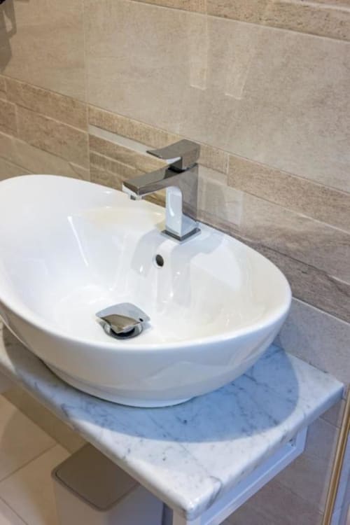 Lavabo rotondo bianco moderno con rubinetto cromato nel bagno La Maison Du Lion, con piano in marmo e parete rivestita in piastrelle beige
