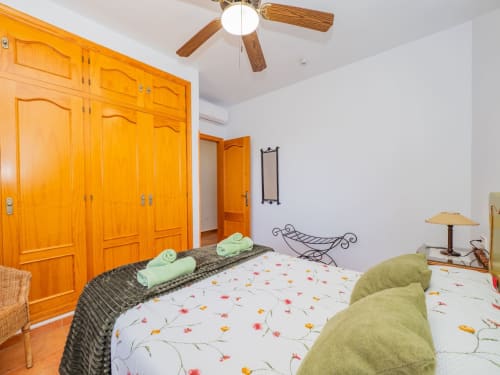 Dormitorio luminoso con armario de madera naranja, ropa de cama estampada, cojines verdes, ventilador de techo y decoración tradicional en Alhaurín