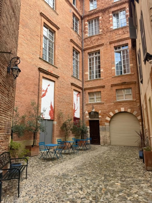 Innenhof des Les Relais Du Capitole in Toulouse mit Backsteingebäuden, blauen Stühlen und gepflastertem Hof
