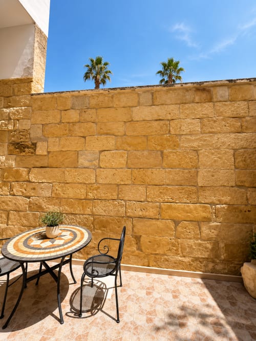 Stone-walled patio at HABITARE Lecce with mosaic table, wrought iron chairs, and palm trees under blue sky