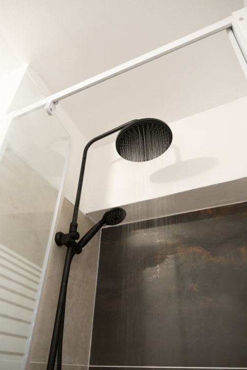 Pommeau de douche moderne noir et douchette à main dans la salle de bains de l'appartement NGAYABE 976 à Mamoudzou, murs carrelés blancs