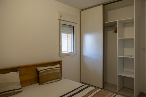 Chambre de l'appartement CP204 à NGAYABE 976, Mamoudzou avec lit en bois, armoire miroir, fenêtre et étagères blanches