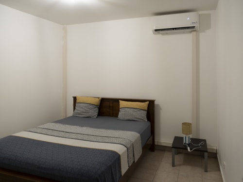 Chambre moderne de l'appartement CP204 à NGAYABE 976, Mamoudzou avec lit en bois foncé, literie grise, oreillers jaunes, climatiseur et