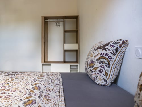 Chambre de l'appartement NGAYABE 976 à Mamoudzou avec literie à motifs, coussin décoratif et armoire en bois.
