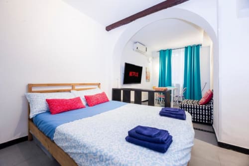 Camera moderna luminosa a La Superba Stay Genova con letto in legno, biancheria blu, cuscini rossi, TV e tende turchesi