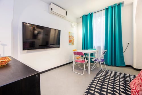 Luminosa zona living di La Superba Stay Genova con TV schermo piatto, tende turchesi, tavolo bianco e sedie colorate
