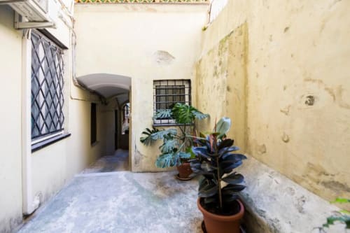 Ingresso di stretto cortile a La Superba Stay Genova con piante in vaso e muri invecchiati, che conduce al palazzo abitativo
