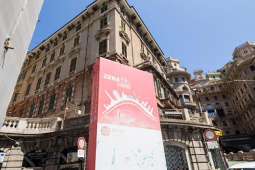 Cartello rosa del bike-sharing ZENA su storica facciata genovese con architettura tradizionale