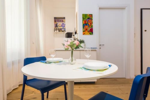 Area dining moderna a La Superba Stay Genova con tavolo rotondo bianco, sedie blu, mise en place e fiori rosa