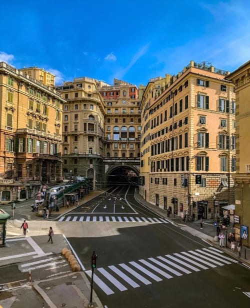 Piazza storica di Genova con edifici rinascimentali gialli, ingresso di tunnel e attraversamenti pedonali sotto cielo azzurro