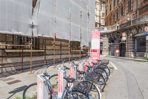 Stazione di bike-sharing in via Genova con biciclette rosse a noleggio parcheggiate davanti a una storica facciata