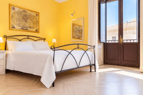 Camera da letto giallo vivace con letto in ferro battuto, biancheria bianca, mappe vintage e balcone con vista su Catania in Sicilia