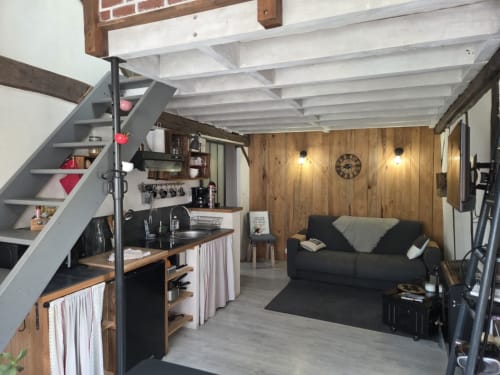 Intérieur de cottage style loft moderne avec kitchenette, canapé noir, accents bois et escalier à La Maréchalerie