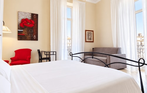 Bright Catania apartment bedroom with red accent chair, wrought iron bed, gray sofa, and city views from floor-to-ceiling