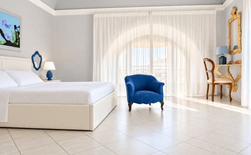 Camera da letto luminosa e moderna nell'appartamento Blue House Catania con letto bianco, poltrona in velluto blu, balcone con vista città e arredamento elegante