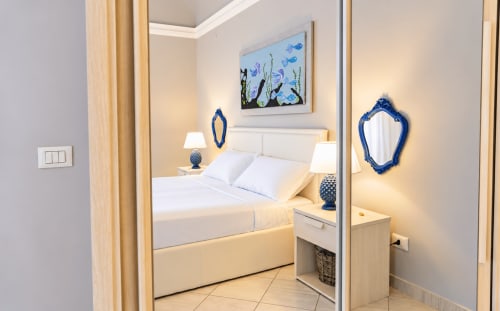 Modern bedroom in Sicily In Home apartment with white bed, blue decorative mirror and artwork, bedside lamps, and light wood
