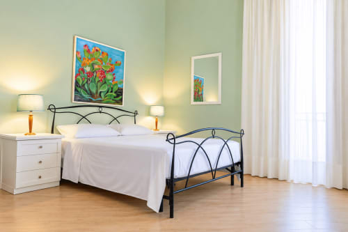Camera da letto luminosa a Sicily In Home con letto in metallo, biancheria bianca, pareti verdi, quadri floreali e pavimento in legno
