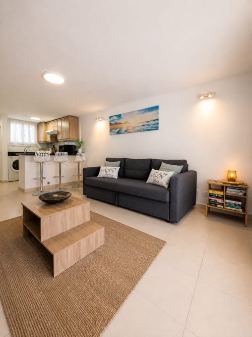 Modern apartment living room with black sofa, wooden coffee table, kitchenette with bar seating, and ocean-view artwork at