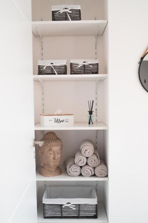 White wall shelving with woven baskets, rolled towels, Buddha head, tissue box, and reed diffuser at Résidence des Sables