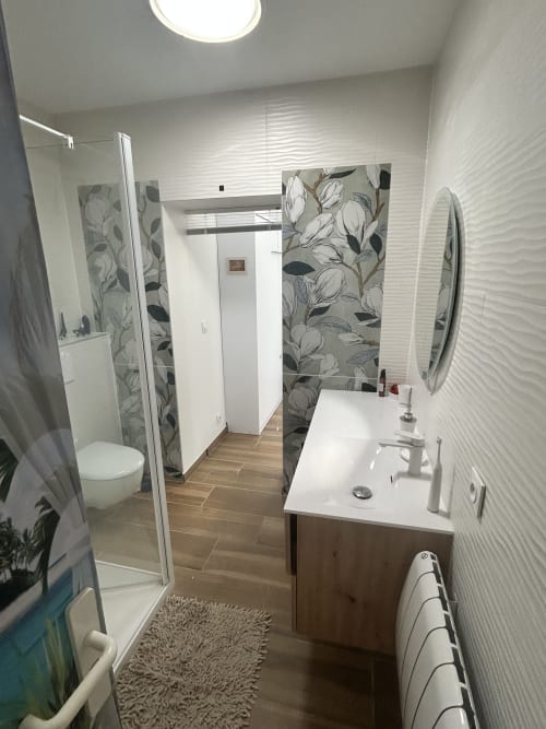 Salle de bains moderne aux carreaux texturés blancs, art mural fleuri, vanité en bois, miroir rond et douche pluie