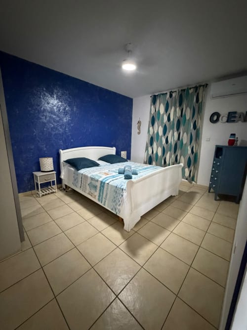 Chambre La Case à Nath avec lit blanc, mur d'accent bleu, rideaux motifs turquoise et sol carrelé au Domaine Des Doudous