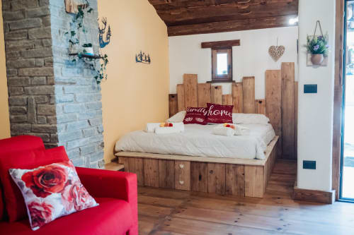 Accogliente camera al Borgo Maira Village di Prazzo con letto in legno, poltrona rossa, muri bianchi e camino in pietra.