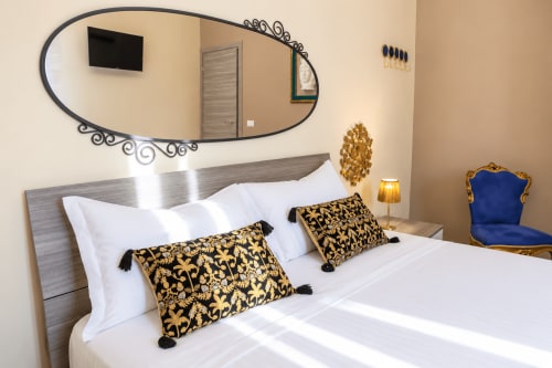 Luxurious bedroom with white bedding, gold and black patterned pillows, ornate mirror, and blue velvet chair in Catania
