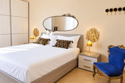 Elegant Catania apartment bedroom with white bed, gold accents, ornate mirror, and blue velvet chair