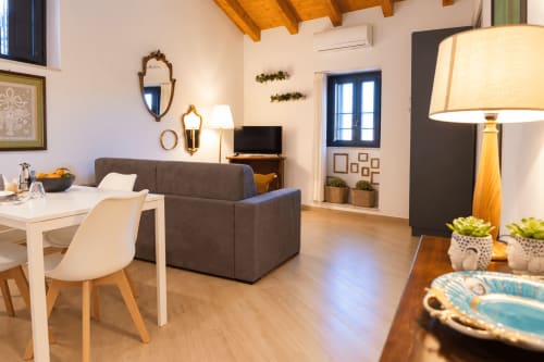 Modern apartment living room in Catania, Sicily with charcoal sofa, dining area, wooden beams, and city views through blue