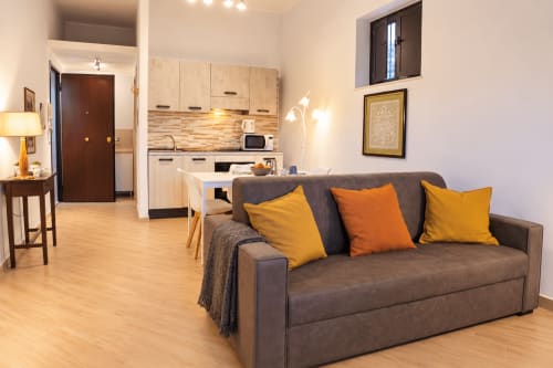 Modern apartment living room with gray sofa, yellow and orange pillows, light wood kitchen, Catania Sicily