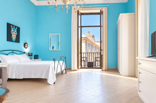 Bright turquoise bedroom in Catania apartment with white bed, balcony overlooking city, modern furnishings and wooden floors