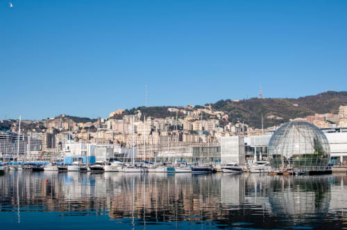 Lungomare di Genova con barche a vela, edifici portuali e paesaggio urbano collinare sotto un cielo blu limpido