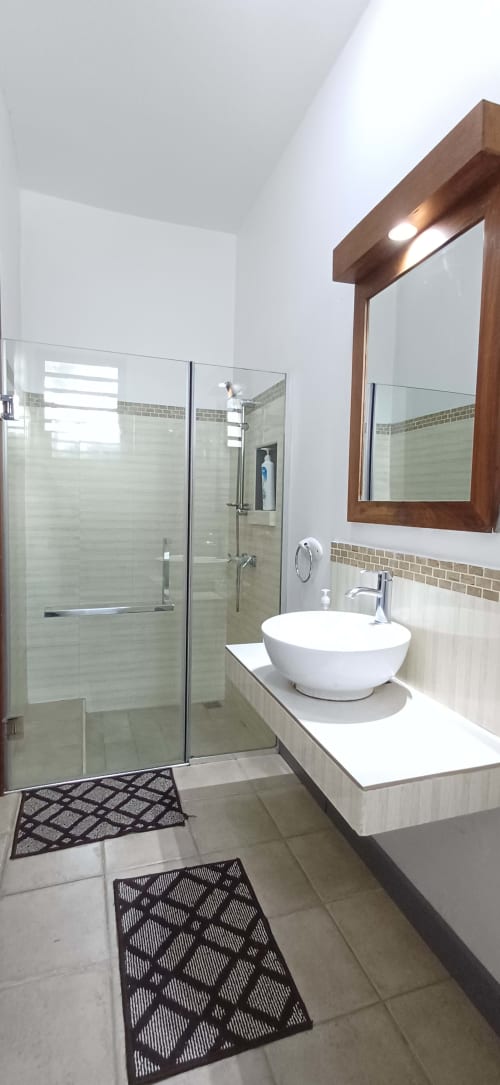 Modern bathroom at Joubarbe Résidence in Moka with vessel sink, glass shower, tiled walls, and warm wooden mirror