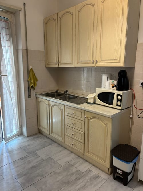 Cucina compatta nell'appartamento di Ostia con mobili color crema, lavello in acciaio inox, microonde e bollitore