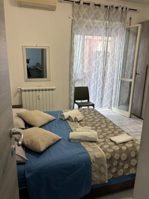 Spaziosa camera da letto a ViOletta Ostia Roma con biancheria blu, plaid con motivo margherite, cuscini, asciugamani arrotolati e accesso al balcone