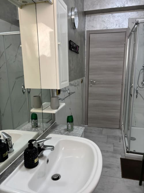 Moderno bagno dell'appartamento ViOletta Ostia Roma con lavabo bianco, mobile con specchio, piastrelle grigie e cabina doccia in vetro