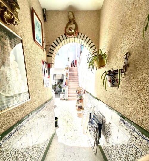 Estrecho pasillo en apartamento de Jerez con azulejos decorativos, plantas colgantes, arte religioso y arco que conduce al patio