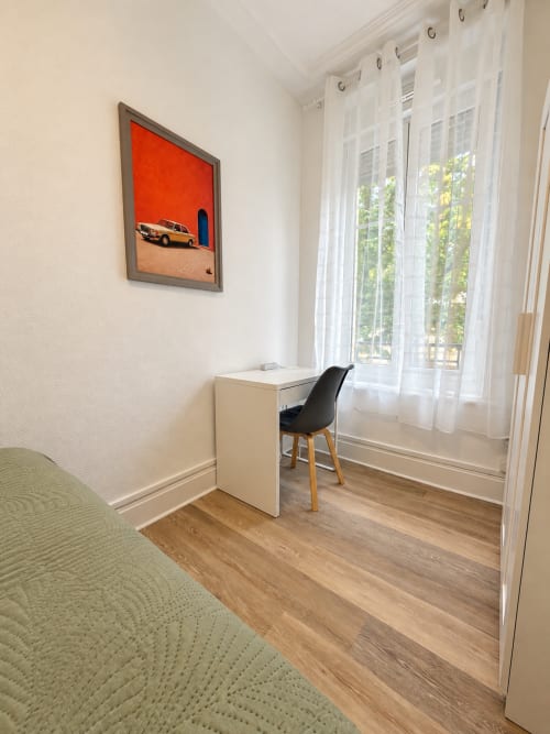 Chambre d'hôtel lumineuse à Nancy-Place avec bureau blanc, chaise noire et tableau encadré rouge près des fenêtres aux voilages délicats