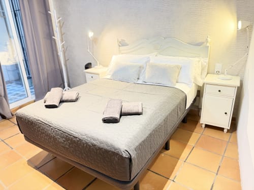Dormitorio moderno en apartamento Stay Gest Valencia con cama de estructura blanca, ropa de cama gris, mesitas blancas, baldosas terracota, y