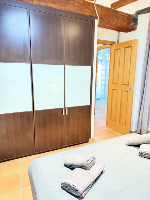 Moderno dormitorio en apartamento de Valencia con armario de madera, toallas dobladas en la cama y puerta de madera hacia la sala