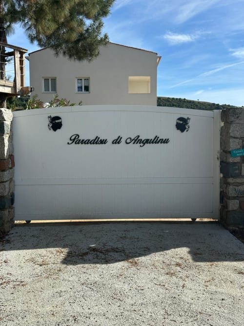 Portail blanc d'entrée à la propriété Paradisu di Anguillnu à Alata, avec maison beige moderne et vue sur les collines verdoyantes