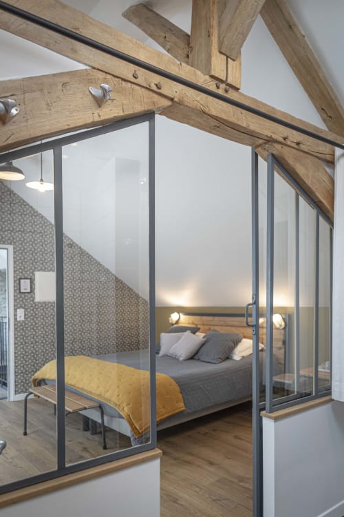 Chambre moderne avec poutres en bois, lit à structure vitrée et linge jaune au gîte Les Olivanies, Monthelon