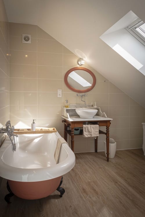 Salle de bain mansardée luxueuse aux Olivanies avec baignoire indépendante, lavabo vasque, miroir ovale, puits de lumière et parquet