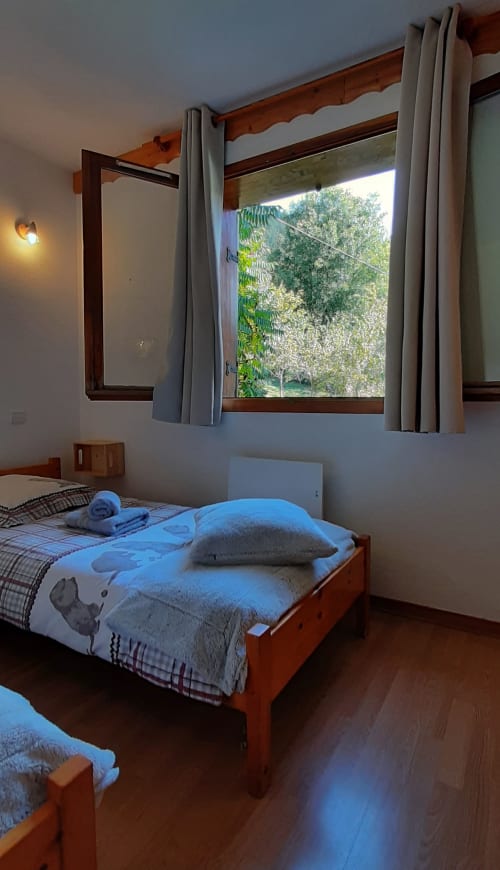 Chambre lumineuse du Chalet De La Yodine avec lit en bois, rideaux gris et vue sur le jardin montagnard par grandes fenêtres