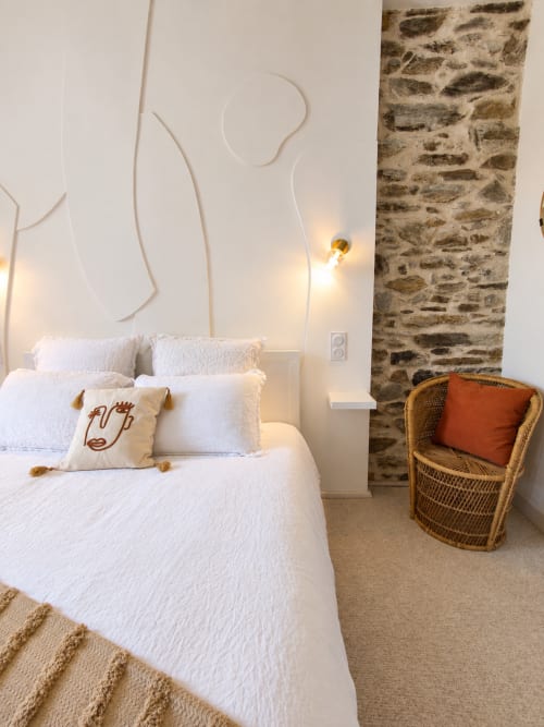 Chambre Aurore lumineuse aux Chambres D'Hespéris, Pleyben avec draps blancs, mur en pierre, fauteuil en osier et éclairage chaleureux