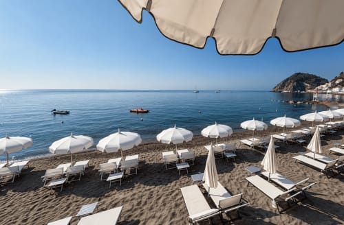 Ombrelloni bianchi e lettini sulla spiaggia sabbiosa del Villa Bina Sea Hotel di Serrara Fontana, con mare blu calmo e barche