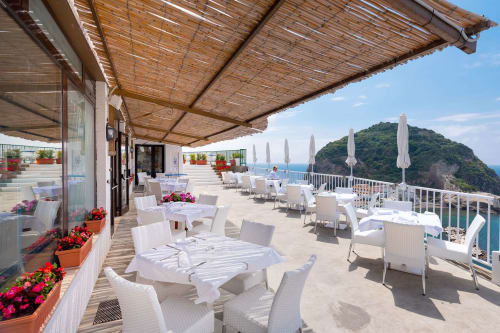 Terrazza dell'Hotel Villa Bina a Serrara Fontana con tavoli da pranzo, sedie bianche, ombrelloni, fiori e vista sul mare
