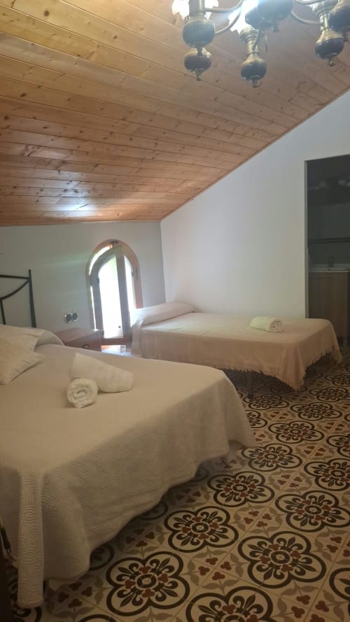 Spacious bedroom at Refugio la Presa with two beds, wooden ceiling, arched window, and decorative tile flooring in Chulilla.
